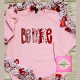 Pink sweatshirt with 'Be Mine' text surrounded by festive tinsel and heart decorations on a wooden surface.